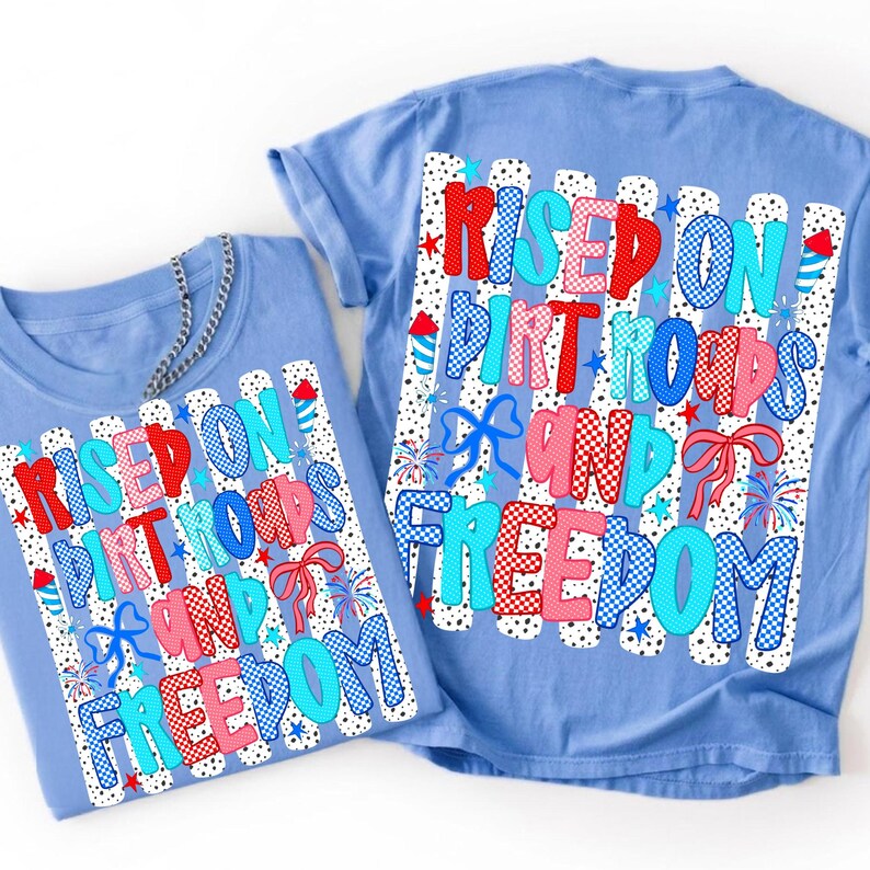 Op de afbeelding: Twee lichtblauwe t-shirts met een grafische print van verticale zwarte en witte strepen. De tekst "RISED ON DIRT ROADS AND FREEDOM" staat in rood, roze en blauw gedessineerde letters, versierd met kleine vuurwerken, sterren en strikjes.