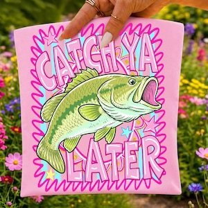 Op de afbeelding: Roze t-shirt met een groen-witte visafbeelding en de tekst "CATCH YA LATER" in roze en blauw. De vis heeft een open mond en is omgeven door sterren en een golvende rand.