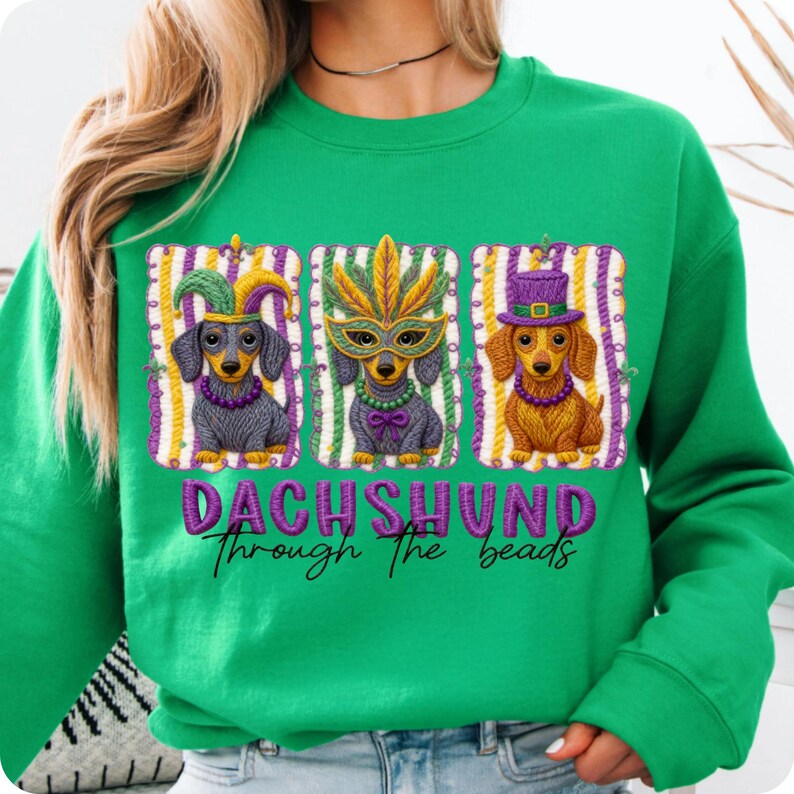 Puede incluir: Sudadera verde con tres ilustraciones de perros salchicha con disfraces de Mardi Gras. Cada perro est&aacute; enmarcado con rayas moradas y doradas. El texto "DACHSHUND through the beads" est&aacute; impreso en morado.