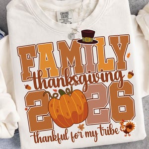 Pode incluir: Uma camisola de gola redonda cor de creme com um gr&aacute;fico que inclui o texto FAMILY THANKSGIVING 2026, thankful for my tribe. O design apresenta um chap&eacute;u de peregrino castanho, uma ab&oacute;bora laranja, bolotas, folhas e um peru.