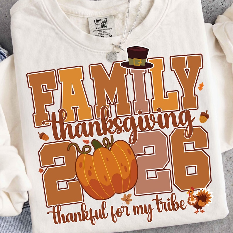 Pode incluir: Uma camisola de gola redonda cor de creme com um gr&aacute;fico que inclui o texto FAMILY THANKSGIVING 2026, thankful for my tribe. O design apresenta um chap&eacute;u de peregrino castanho, uma ab&oacute;bora laranja, bolotas, folhas e um peru.