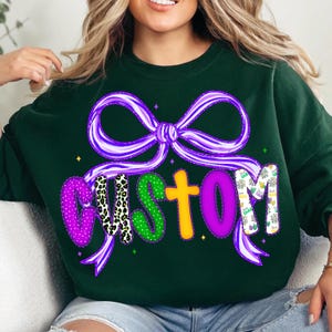 May include: Dark green sweatshirt with a purple bow and the word "CUSTOM" in colorful letters. The letters have various patterns, including leopard print and Mardi Gras designs. The bow and letters are outlined in white.