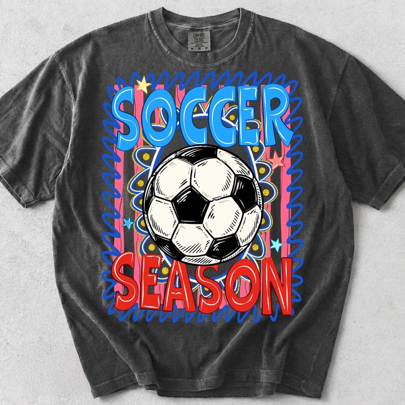 Puede incluir: Camiseta gris oscuro con un gr&aacute;fico de bal&oacute;n de f&uacute;tbol. El dise&ntilde;o presenta las palabras "SOCCER SEASON" en azul y rojo, con un bal&oacute;n de f&uacute;tbol en blanco y negro en el centro. El fondo tiene detalles rosas y amarillos.