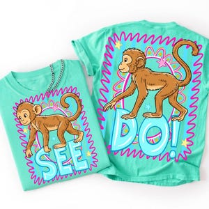 May include: Two turquoise t-shirts with a cartoon monkey design. One shirt says "SEE" and the other says "DO!" in large, light blue letters. The monkey is brown with a long tail, and the design is framed by a pink and blue border.