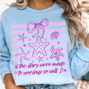 Op de afbeelding: Lichtblauw sweatshirt met een roze ontwerp van zeesterren, strikken en schelpen. De tekst "if the stars were made *To worship so will I!*" is roze gedrukt. Het ontwerp staat op een lichtblauwe achtergrond.