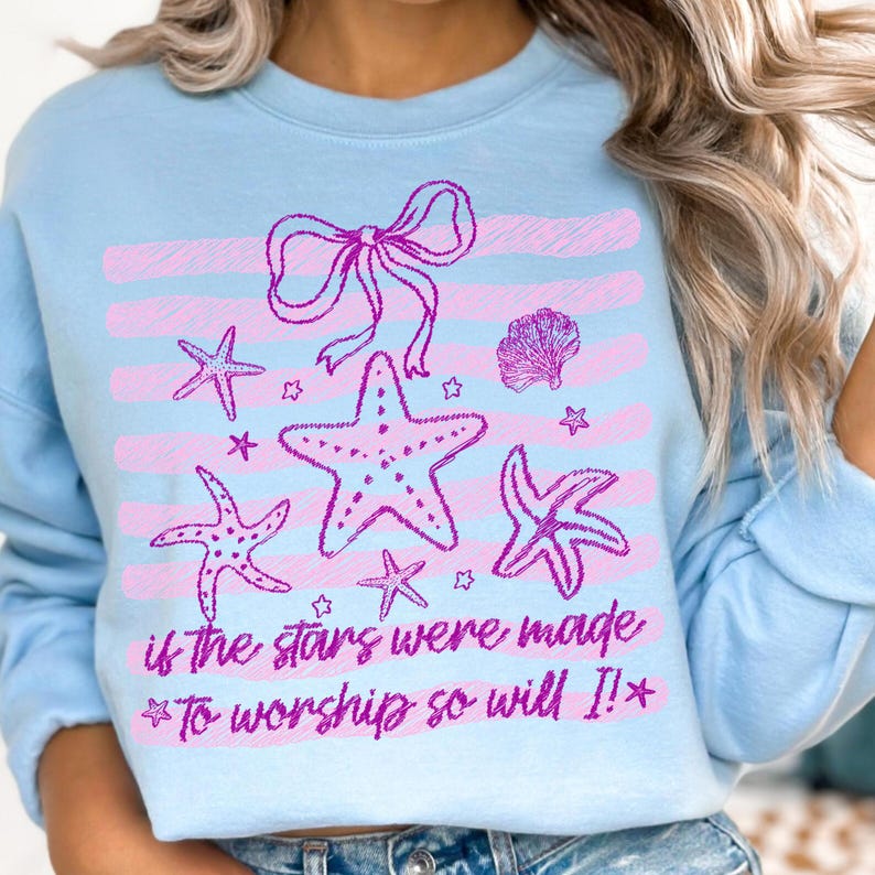 Op de afbeelding: Lichtblauw sweatshirt met een roze ontwerp van zeesterren, strikken en schelpen. De tekst "if the stars were made *To worship so will I!*" is roze gedrukt. Het ontwerp staat op een lichtblauwe achtergrond.