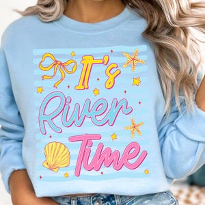 May include: Light blue sweatshirt with the text "It's River Time" in yellow and pink. The design includes a yellow bow, starfish, a seashell, and stars. The sweatshirt is a casual, beach-themed apparel item.
