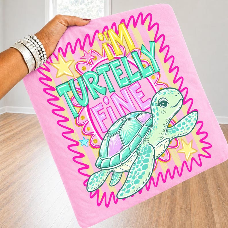 May include: Pink blanket with the text "I'M TURTELLY FINE" and a cartoon turtle illustration. The turtle has a teal shell and is surrounded by a pink and yellow wavy border with stars. The blanket is held up by a person.