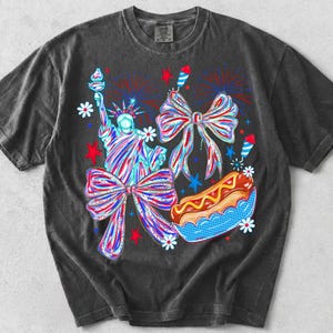 May include: Dark gray t-shirt featuring a colorful graphic design. The design includes the Statue of Liberty, bows, fireworks, a hot dog, and stars, all in red, white, and blue. The shirt has a vintage, washed look.