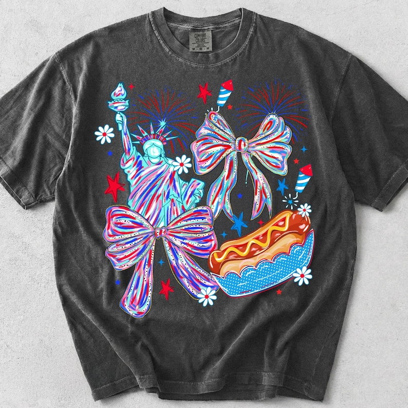 May include: Dark gray t-shirt featuring a colorful graphic design. The design includes the Statue of Liberty, bows, fireworks, a hot dog, and stars, all in red, white, and blue. The shirt has a vintage, washed look.
