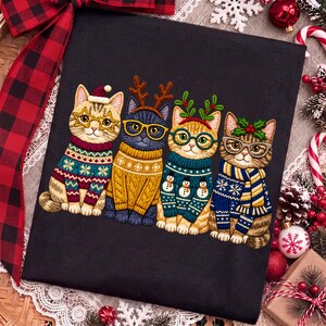 May include: A black t-shirt featuring an embroidered design of four cats wearing holiday sweaters, glasses, and festive accessories like reindeer antlers, a Santa hat, and holly. The cats are depicted in various colors with detailed patterns.
