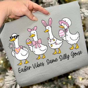 May include: A light gray fabric with a cartoon illustration of four geese dressed for Easter. The geese are white with yellow feet and beaks, wearing accessories like sunglasses, bows, and bunny ears. The text "Easter Vibes Same Silly Goose" is below the geese.