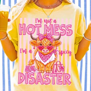 K&ouml;nnte beinhalten: Gelbes T-Shirt mit einer Grafik einer Highland-Kuh mit herzf&ouml;rmiger Sonnenbrille, Schleife und Bandana. Der Text lautet: &bdquo;I'm not a HOT MESS, I'm a spicy DISASTER.&ldquo;