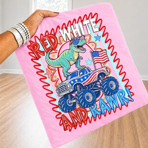 May include: Pink blanket with a cartoon dinosaur wearing sunglasses and riding a monster truck with an American flag. The blanket has the words "RED WHITE AND RAWR!" in a playful font.