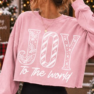 May include: A dusty pink cropped sweatshirt featuring the word JOY in large white letters, with the J decorated with holly leaves, the O with diagonal stripes, and the Y with small crosses. The text To The world is printed below in white.