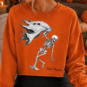 K&ouml;nnte beinhalten: Ein orangefarbenes Langarm-Sweatshirt mit einem Grafikdruck eines Skeletts, das ein wei&szlig;es Laken mit einem Sternenhimmel-Muster h&auml;lt. Der Text "Sheet Happens.." ist in Schwarz unter dem Skelett auf der Vorderseite des Kleidungsst&uuml;cks gedruckt.