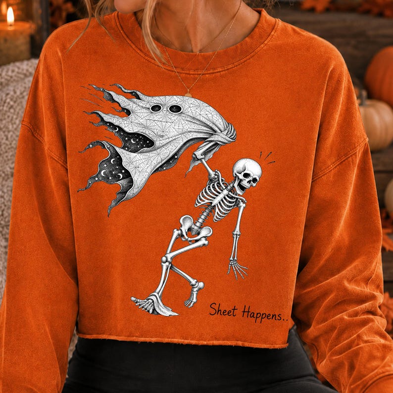 K&ouml;nnte beinhalten: Ein orangefarbenes Langarm-Sweatshirt mit einem Grafikdruck eines Skeletts, das ein wei&szlig;es Laken mit einem Sternenhimmel-Muster h&auml;lt. Der Text "Sheet Happens.." ist in Schwarz unter dem Skelett auf der Vorderseite des Kleidungsst&uuml;cks gedruckt.