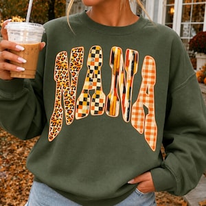 May include: An olive green crewneck sweatshirt featuring the word NANA printed in large, stylized letters. Each letter has a unique pattern: leopard print, black and orange checkerboard, vertical orange and black brushstrokes, and orange gingham.
