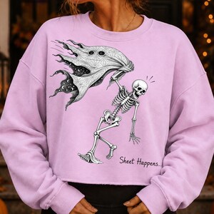 K&ouml;nnte beinhalten: Ein hellviolettes Langarm-Sweatshirt mit einer schwarz-wei&szlig;en Grafik eines Skeletts, das ein Geisterlaken h&auml;lt, welches ein Sternenhimmel-Muster im Inneren zeigt. Der Text "Sheet Happens.." ist unten rechts in Schwarz aufgedruckt.