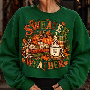 Puede incluir: Una sudadera verde oscuro de cuello redondo con un estampado gr&aacute;fico que incluye el texto "SWEATER WEATHER". El dise&ntilde;o muestra una calabaza naranja grande, una pila de mantas de punto dobladas, una taza humeante "GOOD THINGS AHEAD" y bellotas.