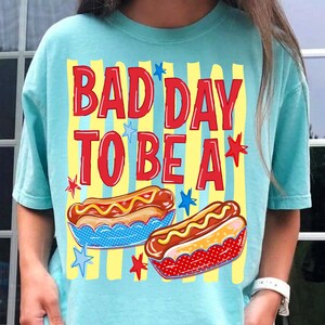 K&ouml;nnte beinhalten: Hellblaues T-Shirt mit rotem Schriftzug "BAD DAY TO BE A". Das Design zeigt zwei Hotdogs mit gelbem Senf und Sternen. Der Hintergrund hat gelbe vertikale Streifen.