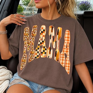 May include: A dark brown short-sleeve t-shirt featuring the word MAMA printed in large, stylized letters. Each letter has a distinct pattern: leopard print, black and orange checkered, vertical brush strokes, and orange and white gingham.