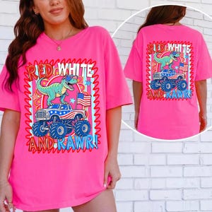 May include: A bright pink t-shirt with a graphic design featuring a green dinosaur wearing sunglasses atop a monster truck with an American flag. The text reads "RED WHITE AND RAWR!" in a wavy, red and blue frame.