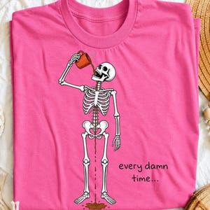 May include: A bright pink t-shirt featuring a graphic of a white skeleton holding an orange mug. The skeleton is pouring a dark brown liquid from the mug into its mouth, which flows through its ribcage and out onto the floor. The text reads every damn time...