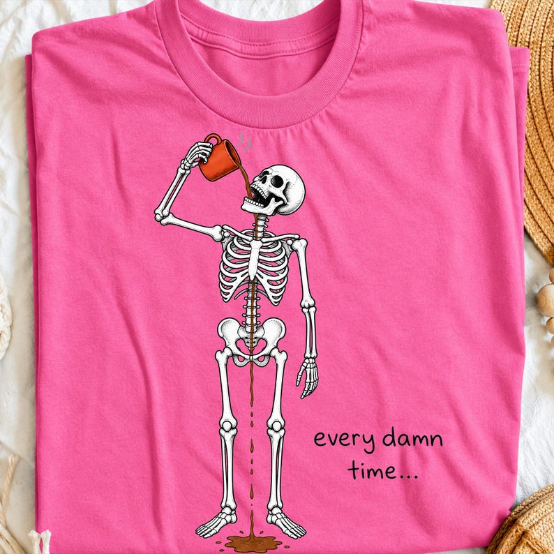 May include: A bright pink t-shirt featuring a graphic of a white skeleton holding an orange mug. The skeleton is pouring a dark brown liquid from the mug into its mouth, which flows through its ribcage and out onto the floor. The text reads every damn time...