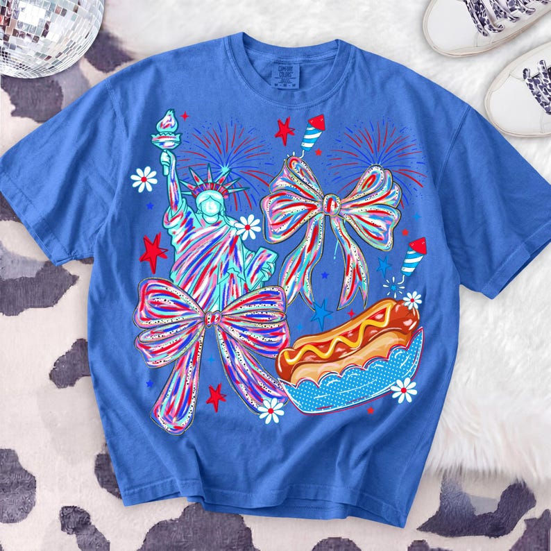 May include: Blue t-shirt with a patriotic design featuring the Statue of Liberty, fireworks, bows, a hot dog, and stars. The graphic is in red, white, and blue colors, with white daisy accents. The shirt is a comfortable, casual style.