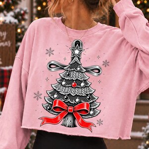 K&ouml;nnte beinhalten: Ein hellrosa, lang&auml;rmeliges bauchfreies Sweatshirt mit einer grafischen Illustration eines stilisierten Weihnachtsbaums. Der Baum ist mit schwarz-wei&szlig;en Ornamenten, einer leuchtend roten Schleife an der Basis und einem Stern an der Spitze verziert.