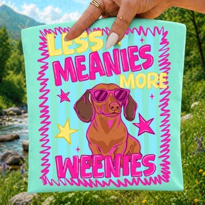 May include: A turquoise tote bag with the text "LESS MEANIES MORE WEENIES" in yellow and pink. The bag features a cartoon dachshund wearing pink sunglasses. The design includes stars and a wavy pink border.