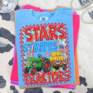 Pode incluir: Camiseta azul clara com as palavras "STARS STRIPES TRACTORS" em vermelho e azul. O gr&aacute;fico apresenta um trator verde puxando uma carro&ccedil;a de fardos de feno e uma bandeira americana. A camiseta est&aacute; dobrada sobre outras camisetas coloridas.