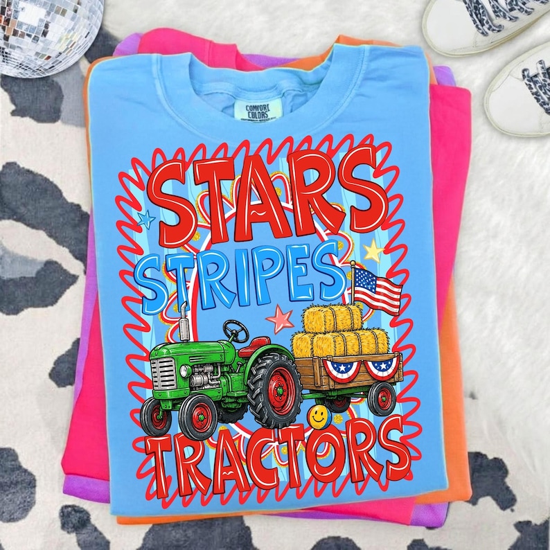 Pode incluir: Camiseta azul clara com as palavras "STARS STRIPES TRACTORS" em vermelho e azul. O gr&aacute;fico apresenta um trator verde puxando uma carro&ccedil;a de fardos de feno e uma bandeira americana. A camiseta est&aacute; dobrada sobre outras camisetas coloridas.