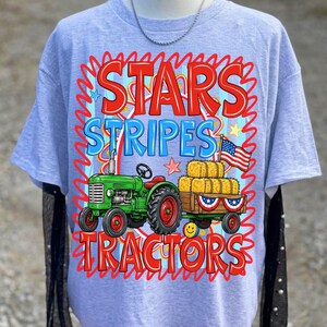 Pode incluir: Camiseta azul clara com um gr&aacute;fico de um trator verde puxando uma carro&ccedil;a de fardos de feno e uma bandeira americana. O texto "STARS STRIPES TRACTORS" est&aacute; em vermelho e azul.