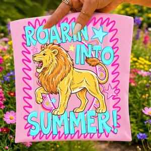 May include: Pink graphic tee with a cartoon lion illustration. The lion is yellow with an orange mane and tail. Text in turquoise reads "ROAR INTO SUMMER!" with a pink wavy border and stars.