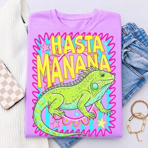 K&ouml;nnte beinhalten: Ein lila T-Shirt mit einer gr&uuml;nen Leguan-Illustration und dem Text "HASTA MANANA" in Gelb. Das Design umfasst rosa, blaue und gelbe Akzente. Das T-Shirt wird mit einer blauen Jeans und einem goldenen Armband pr&auml;sentiert.