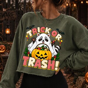 May include: An olive green long-sleeve cropped sweatshirt featuring a graphic of a raccoon dressed as a ghost holding a jack-o'-lantern bucket filled with candy. The text reads TRICK OR TRASH in a red and white checkered pattern with yellow outlines.