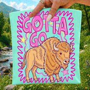 May include: A light blue tote bag with the words "GOTTA GO" in pink bubble letters, a cartoon bison, and a pink zig-zag border. The bison is tan with brown details. The bag is held by a hand with gold rings.