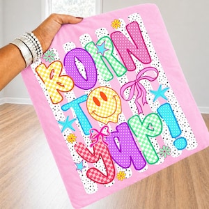 May include: Pink blanket featuring the phrase "BORN TO YAP!" in vibrant bubble letters. The design includes a striped background with polka dots, bows, stars, and floral accents for a playful look.