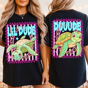 May include: Black t-shirt with a cartoon turtle design. The front says "Lil Dude" and the back says "Duuude". The turtle is green and yellow, framed by a pink and blue decorative border.