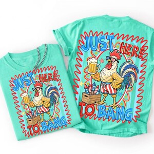 May include: Two turquoise t-shirts with a rooster graphic. The rooster wears sunglasses and patriotic shorts, holding a beer and standing near fireworks. Text reads "JUST HERE TO BANG" in a red and blue design.