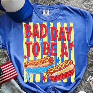 K&ouml;nnte beinhalten: Blaues T-Shirt mit dem roten Text "BAD DAY TO BE A". Das Design umfasst gelbe Streifen, Sterne und Abbildungen von Hotdogs. Eine blau-wei&szlig;e Baseballkappe und eine amerikanische Flagge sind ebenfalls zu sehen.