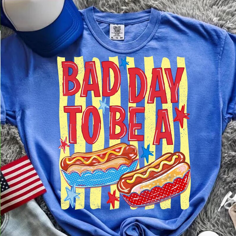 K&ouml;nnte beinhalten: Blaues T-Shirt mit dem roten Text "BAD DAY TO BE A". Das Design umfasst gelbe Streifen, Sterne und Abbildungen von Hotdogs. Eine blau-wei&szlig;e Baseballkappe und eine amerikanische Flagge sind ebenfalls zu sehen.