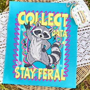 May include: A turquoise tea towel featuring a cartoon raccoon with a rock-and-roll hand gesture. The text "COLLECT DATA STAY FERAL" is displayed in yellow and pink. The design includes stars and lightning bolts. The towel is made of fabric.