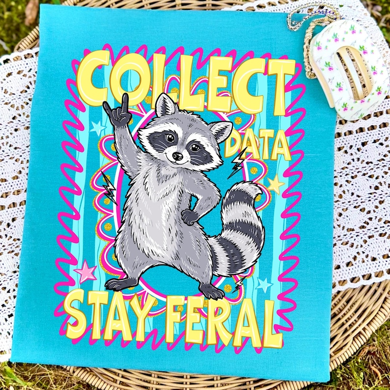 May include: A turquoise tea towel featuring a cartoon raccoon with a rock-and-roll hand gesture. The text "COLLECT DATA STAY FERAL" is displayed in yellow and pink. The design includes stars and lightning bolts. The towel is made of fabric.