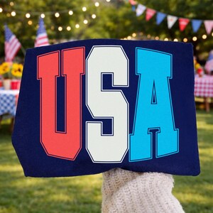 May include: A navy blue sign with the word "USA" in bold, block letters. The letters are outlined in white, with the "U" in red, the "S" in white, and the "A" in blue. The sign is held up outdoors, with a blurred background of a backyard.