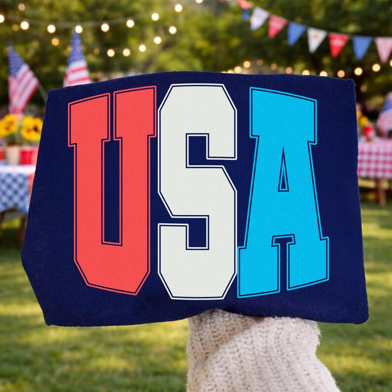 May include: A navy blue sign with the word "USA" in bold, block letters. The letters are outlined in white, with the "U" in red, the "S" in white, and the "A" in blue. The sign is held up outdoors, with a blurred background of a backyard.