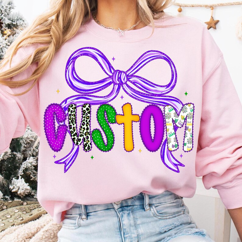 May include: A light pink sweatshirt with a large purple bow design and the word "CUSTOM" in colorful, patterned letters. The letters are decorated with polka dots, animal print, and Mardi Gras-themed patterns. Green and gold stars are scattered around the design.