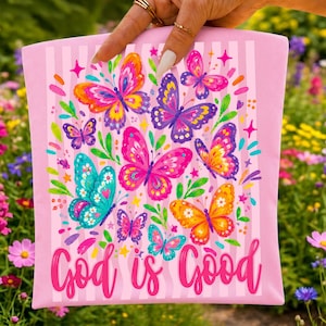 May include: A light pink rectangular fabric item featuring a vertical striped pattern and a colorful print of various butterflies in shades of pink, purple, blue, orange, and yellow, with the text God is Good written in a pink cursive script.